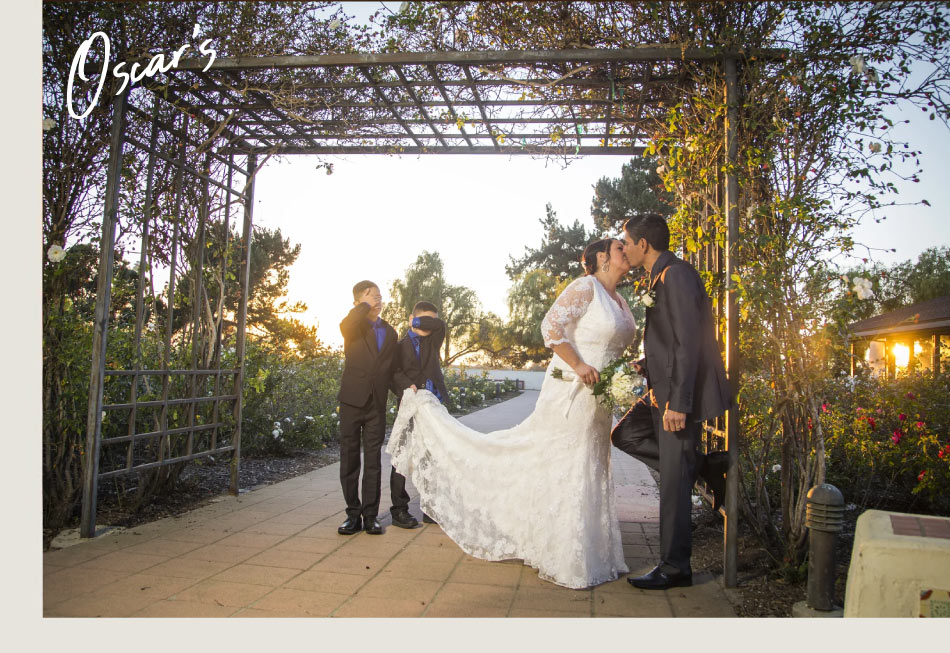 Bride and groom kissing under a garden arbor at golden hour while two young ring bearers hold her dress train, wedding photography