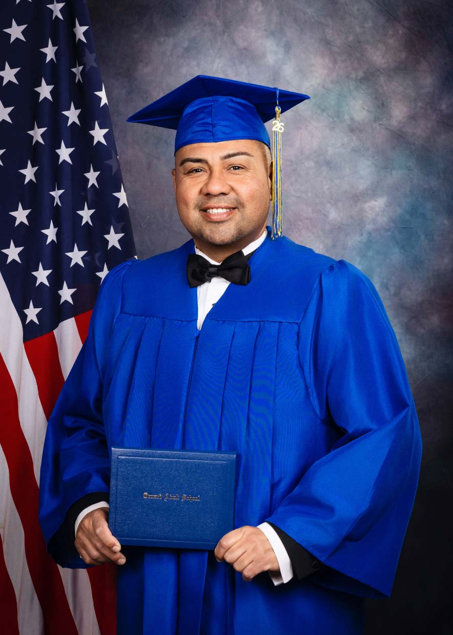 Graduation portrait of a man in blue cap and gown with bow tie holding his diploma, studio backdrop with American flag