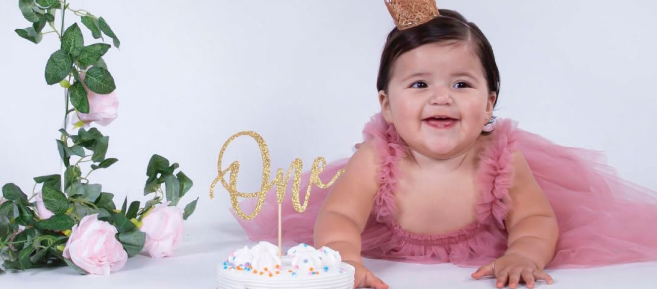 Baby girl in pink tulle dress and gold crown smiling behind a smash cake for her first birthday, studio photoshoot