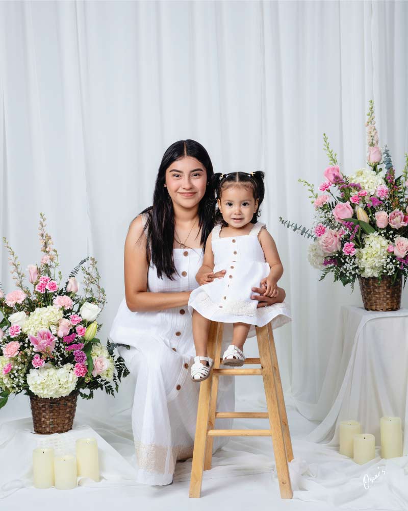 Mother and toddler daughter in matching white button-front sundresses, the toddler seated on a wooden stool, framed by pink flower baskets and candles.