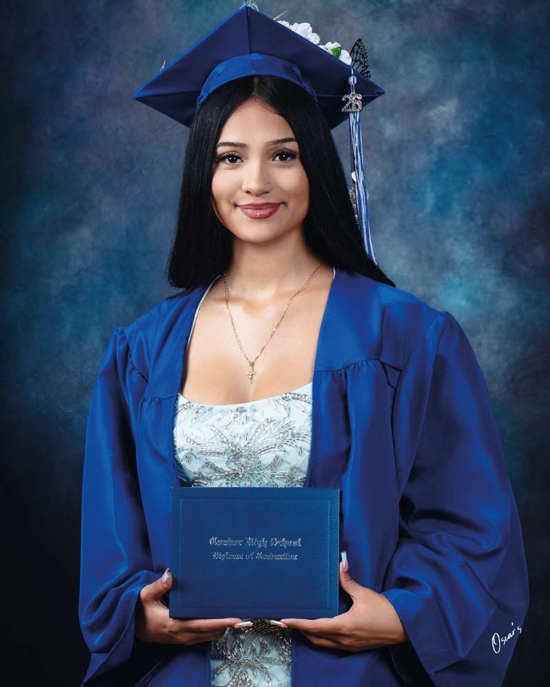 Graduate with long black hair in a blue gown and a flower-decorated cap with a "26" tassel charm, holding a Condor High School diploma.