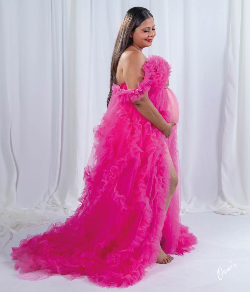 A full-length portrait of a smiling pregnant woman in a dramatic, long-trained off-the-shoulder pink ruffled dress posing sideways in a studio.