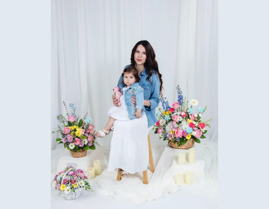 Mother in a denim shirt and white skirt holding her toddler daughter in a matching denim jacket on a stool, framed by colorful spring flower baskets and candles.
