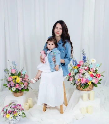Mother in a denim shirt and white skirt holding her toddler daughter in a matching denim jacket on a stool, framed by colorful spring flower baskets and candles.