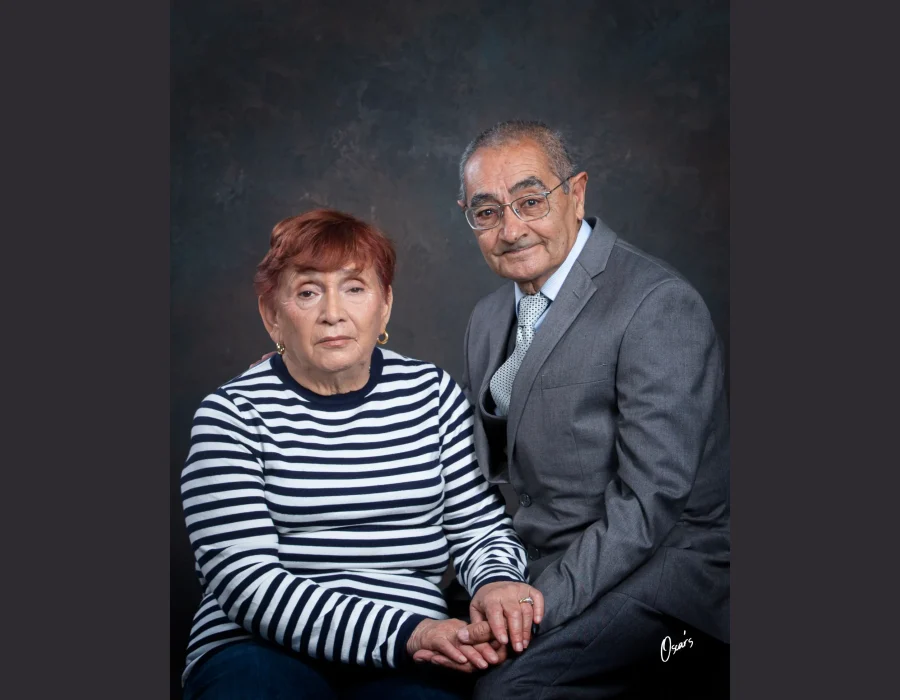 Studio portrait of an elderly couple holding hands, the wife in a navy and white striped sweater and the husband in a gray suit and glasses, at Oscar's Photography and Video.