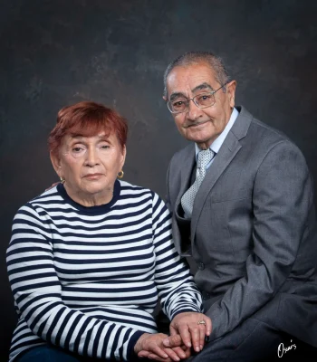 Studio portrait of an elderly couple holding hands, the wife in a navy and white striped sweater and the husband in a gray suit and glasses, at Oscar's Photography and Video.
