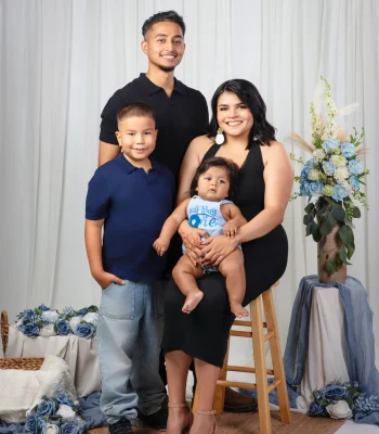 Young family portrait of a father, son, and mother holding their baby boy in a "Half Way to One" romper, styled with dusty blue roses and a white curtain backdrop.
