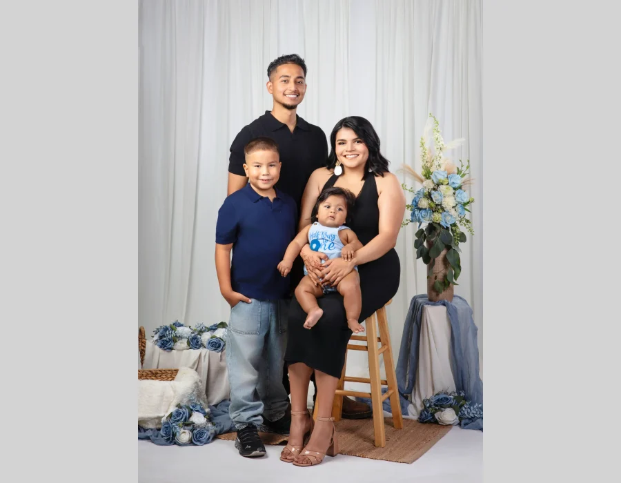 Young family portrait of a father, son, and mother holding their baby boy in a "Half Way to One" romper, styled with dusty blue roses and a white curtain backdrop.