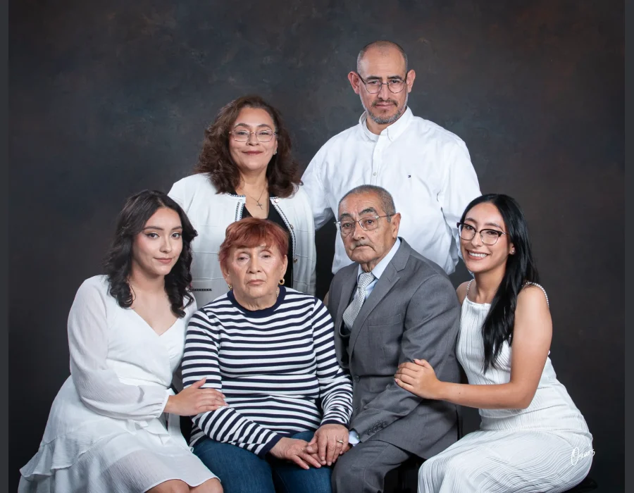 Three-generation family portrait of grandparents seated at center, their adult children standing behind, and two granddaughters in white dresses on either side.