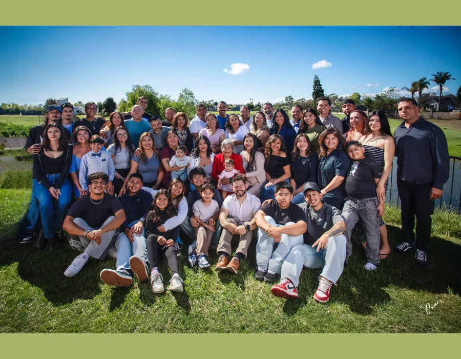 Large multigenerational family reunion portrait of dozens of relatives gathered on a grassy hill under a bright blue sky, with the grandmother in red at the center.