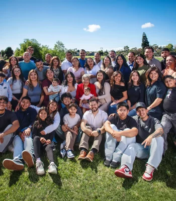 Large multigenerational family reunion portrait of dozens of relatives gathered on a grassy hill under a bright blue sky, with the grandmother in red at the center.