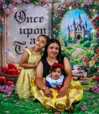 Mother with her two daughters on a storybook-themed set with a castle and "Once Upon a Time" book backdrop, the baby in a yellow princess dress and the older girl in yellow lace.