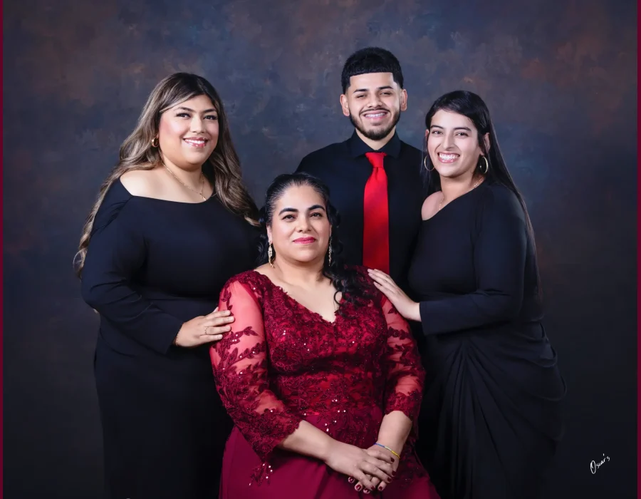 Portrait of a mother in a burgundy lace gown seated in front of her two adult daughters in black dresses and her son in a black shirt and red tie.