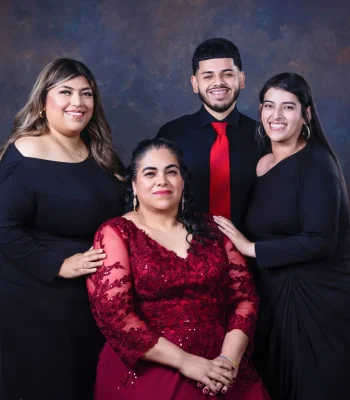Portrait of a mother in a burgundy lace gown seated in front of her two adult daughters in black dresses and her son in a black shirt and red tie.
