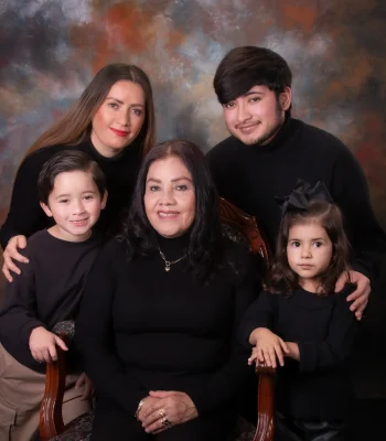 Family portrait of a grandmother seated in a carved wooden chair, surrounded by her daughter, grandson, and two young grandchildren, all dressed in black.