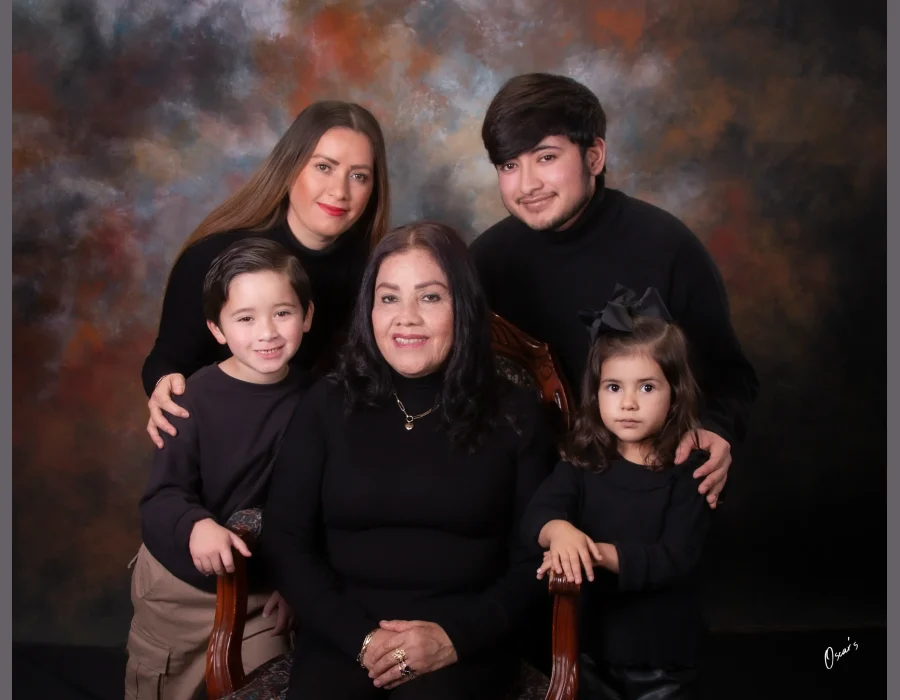 Family portrait of a grandmother seated in a carved wooden chair, surrounded by her daughter, grandson, and two young grandchildren, all dressed in black.