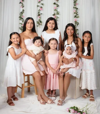 Vertical crop of the pastel family portrait showing seven girls and two babies in pink and white dresses among hanging rose vines and white drapery.