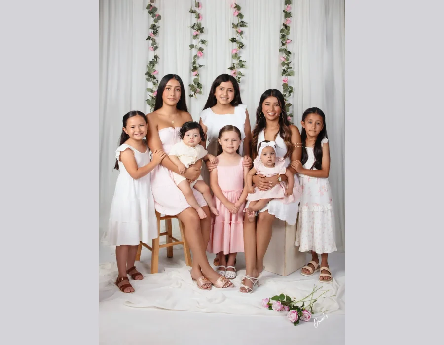 Vertical crop of the pastel family portrait showing seven girls and two babies in pink and white dresses among hanging rose vines and white drapery.