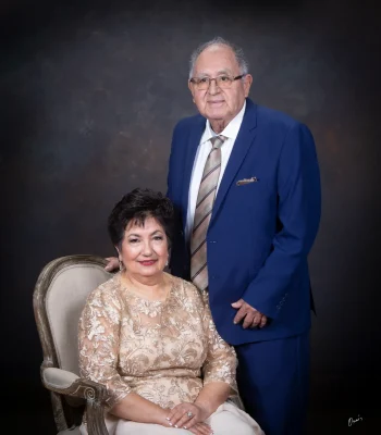 Elegant studio portrait of an older couple, the wife seated in a gold lace dress on a carved armchair and the husband standing beside her in a royal blue suit and striped tie.