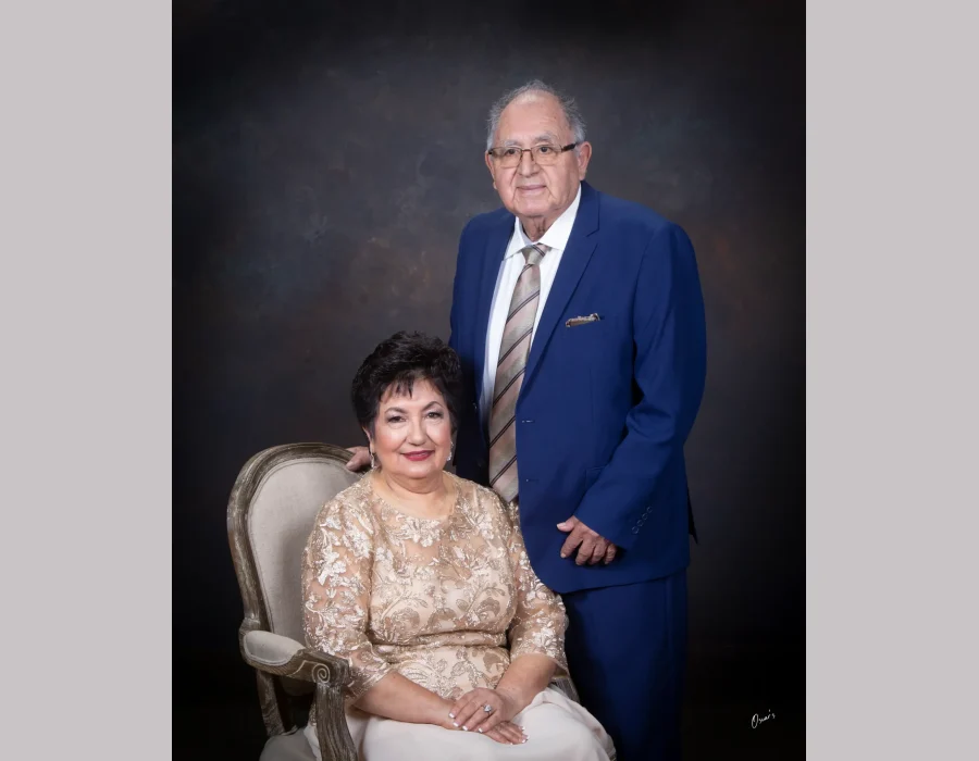 Elegant studio portrait of an older couple, the wife seated in a gold lace dress on a carved armchair and the husband standing beside her in a royal blue suit and striped tie.