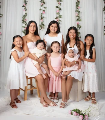 Studio family portrait of seven girls and two babies in coordinated pink and white dresses, posed before a white curtain draped with pink rose vines at Oscar's Photography and Video.