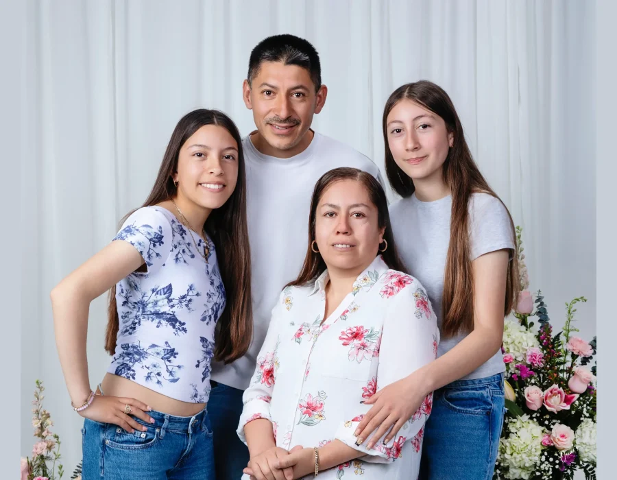 Family portrait of a father in a white tee, a mother in a pink floral blouse, and two teen daughters in casual tops and jeans, posed before a white curtain with pink flower arrangements.