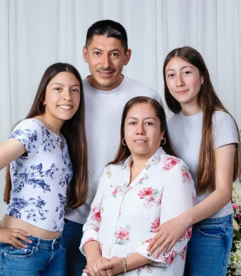 Family portrait of a father in a white tee, a mother in a pink floral blouse, and two teen daughters in casual tops and jeans, posed before a white curtain with pink flower arrangements.
