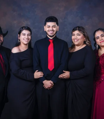 Formal family portrait of a father in a black suit, cowboy hat, and red tie, a son in a black shirt and red tie, two daughters in black dresses, and a mother in a burgundy lace gown.