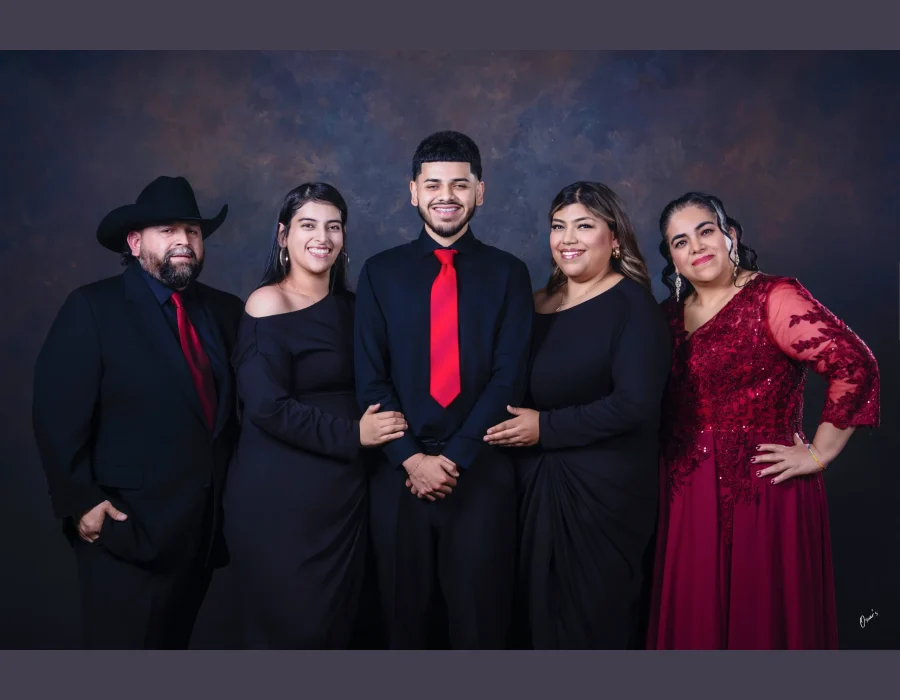 Formal family portrait of a father in a black suit, cowboy hat, and red tie, a son in a black shirt and red tie, two daughters in black dresses, and a mother in a burgundy lace gown.
