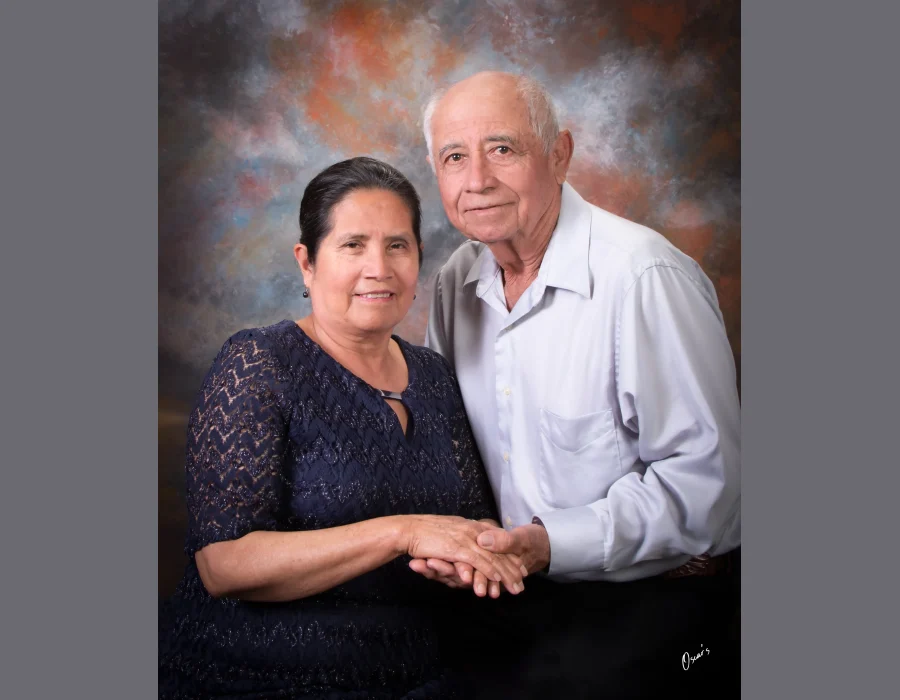 Studio portrait of an older couple holding hands, the wife in a navy lace dress and the husband in a light gray button-up shirt, against a painted backdrop at Oscar's Photography and Video.