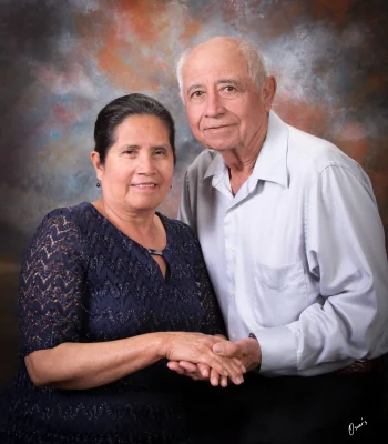 Studio portrait of an older couple holding hands, the wife in a navy lace dress and the husband in a light gray button-up shirt, against a painted backdrop at Oscar's Photography and Video.