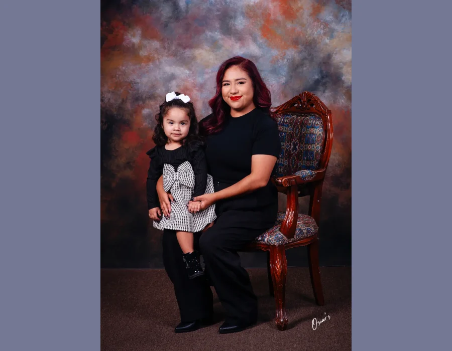 Mother with burgundy-red hair seated in a carved wooden armchair beside her toddler daughter in a black and white houndstooth dress with a white hair bow.