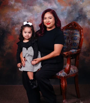 Mother with burgundy-red hair seated in a carved wooden armchair beside her toddler daughter in a black and white houndstooth dress with a white hair bow.