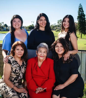 Outdoor multigenerational portrait of five women, with the grandmother in a red suit seated at center, posed by a lake on a sunny day.