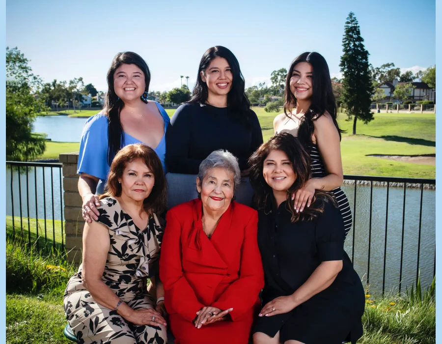 Outdoor multigenerational portrait of five women, with the grandmother in a red suit seated at center, posed by a lake on a sunny day.