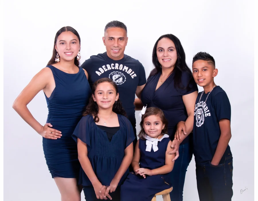 Family of six in coordinated navy outfits, including parents, a teen daughter, a son, and two young daughters, posed against a bright white studio backdrop.