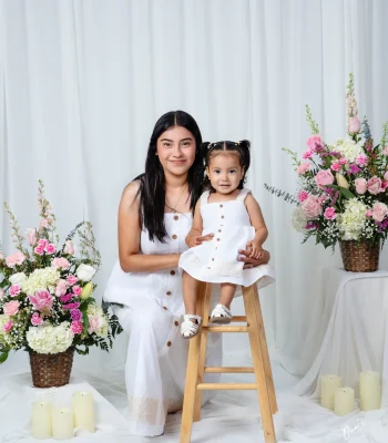 Mother and toddler daughter in matching white button-front sundresses, the toddler seated on a wooden stool, framed by pink flower baskets and candles.