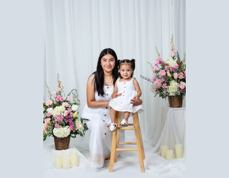 Mother and toddler daughter in matching white button-front sundresses, the toddler seated on a wooden stool, framed by pink flower baskets and candles.