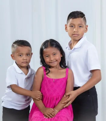 Studio portrait of three siblings, two brothers in white polo shirts on either side of their smiling sister in a bright pink sundress, against a soft white curtain.