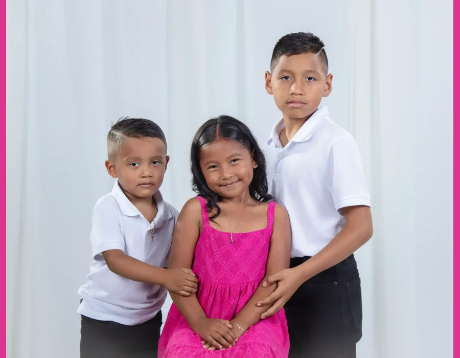 Studio portrait of three siblings, two brothers in white polo shirts on either side of their smiling sister in a bright pink sundress, against a soft white curtain.
