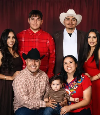 Western-style family portrait of parents holding their baby, surrounded by two adult daughters and two sons, one in a cream cowboy hat, before a deep red curtain at Oscar's Photography and Video.