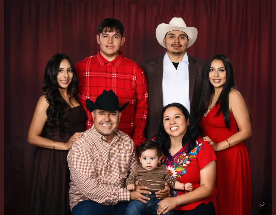 Western-style family portrait of parents holding their baby, surrounded by two adult daughters and two sons, one in a cream cowboy hat, before a deep red curtain at Oscar's Photography and Video.