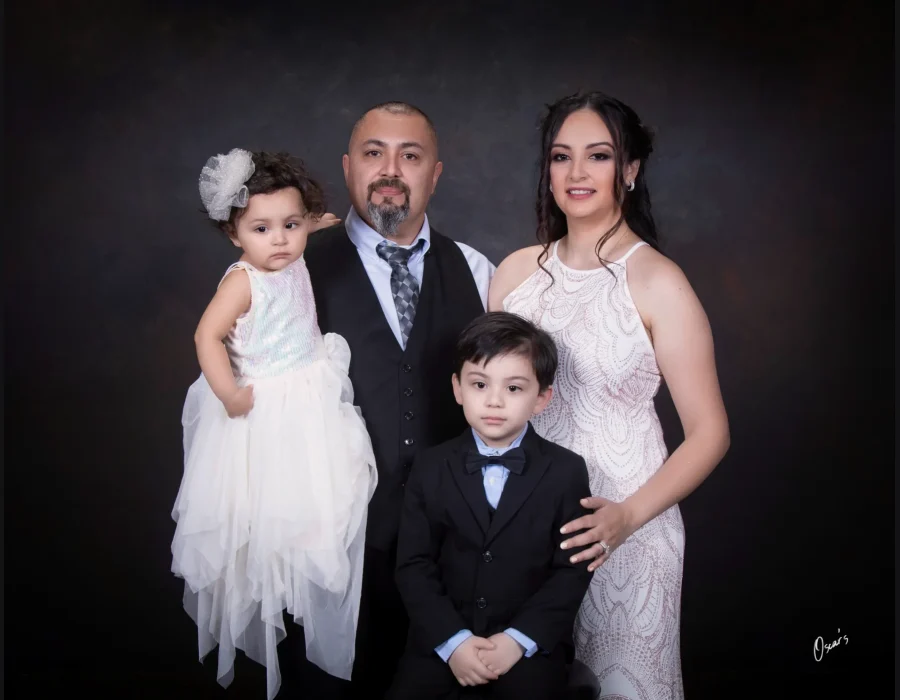 Formal family portrait of a father in a vest and tie, a mother in a sparkling white gown, a toddler daughter in a tulle dress, and a young son in a black suit and bow tie.