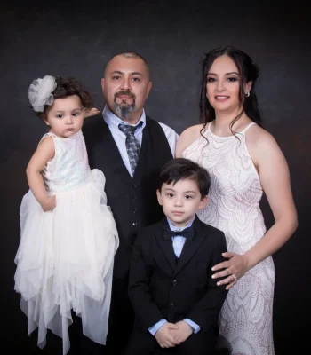 Formal family portrait of a father in a vest and tie, a mother in a sparkling white gown, a toddler daughter in a tulle dress, and a young son in a black suit and bow tie.