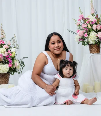 Mother in a white sundress sitting on the floor beside her baby daughter in a white dress with a strawberry appliqué, flanked by pink and white flower baskets.