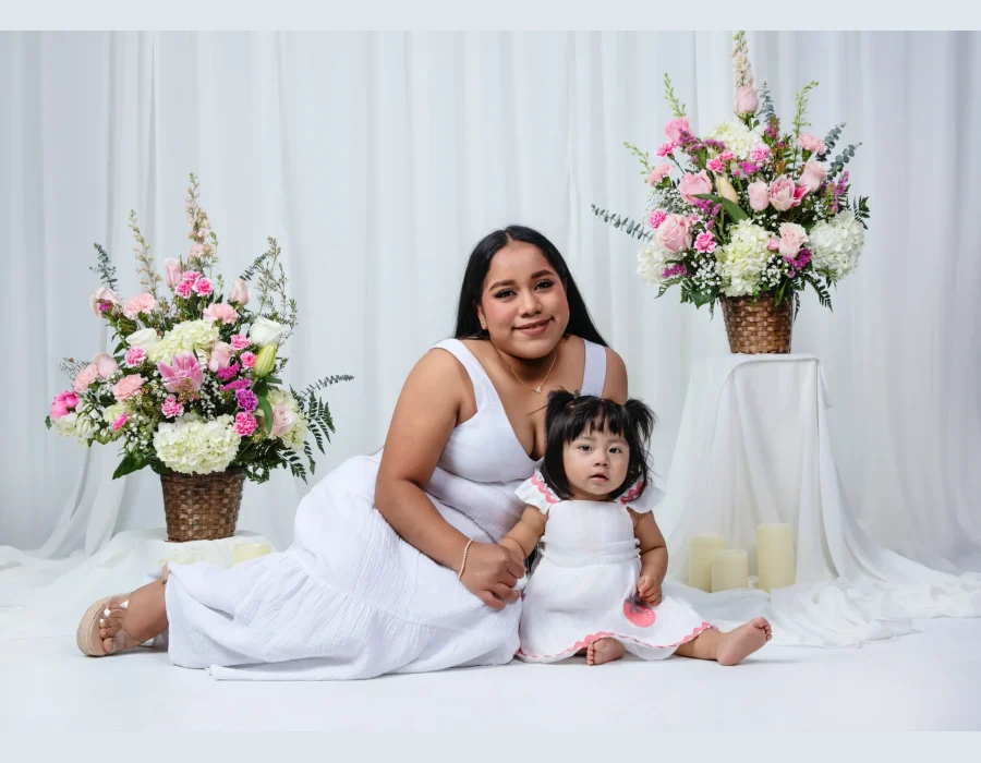 Mother in a white sundress sitting on the floor beside her baby daughter in a white dress with a strawberry appliqué, flanked by pink and white flower baskets.