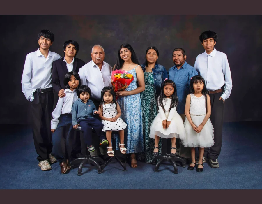 Extended family portrait of twelve, including a grandfather, parents, and children of all ages, gathered around a smiling young woman holding a colorful bouquet.
