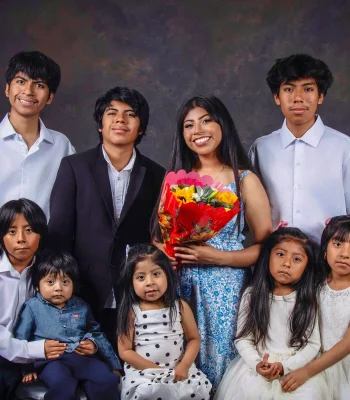 Nine siblings posed together in white shirts and dresses around their older sister, who holds a bright bouquet and wears a blue floral sundress.