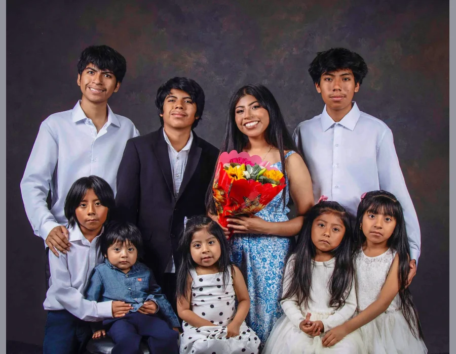 Nine siblings posed together in white shirts and dresses around their older sister, who holds a bright bouquet and wears a blue floral sundress.