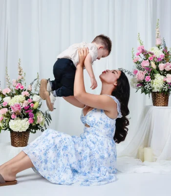 Playful portrait of a mother in a blue floral sundress sitting on the floor and lifting her toddler son overhead, surrounded by pink flower arrangements.